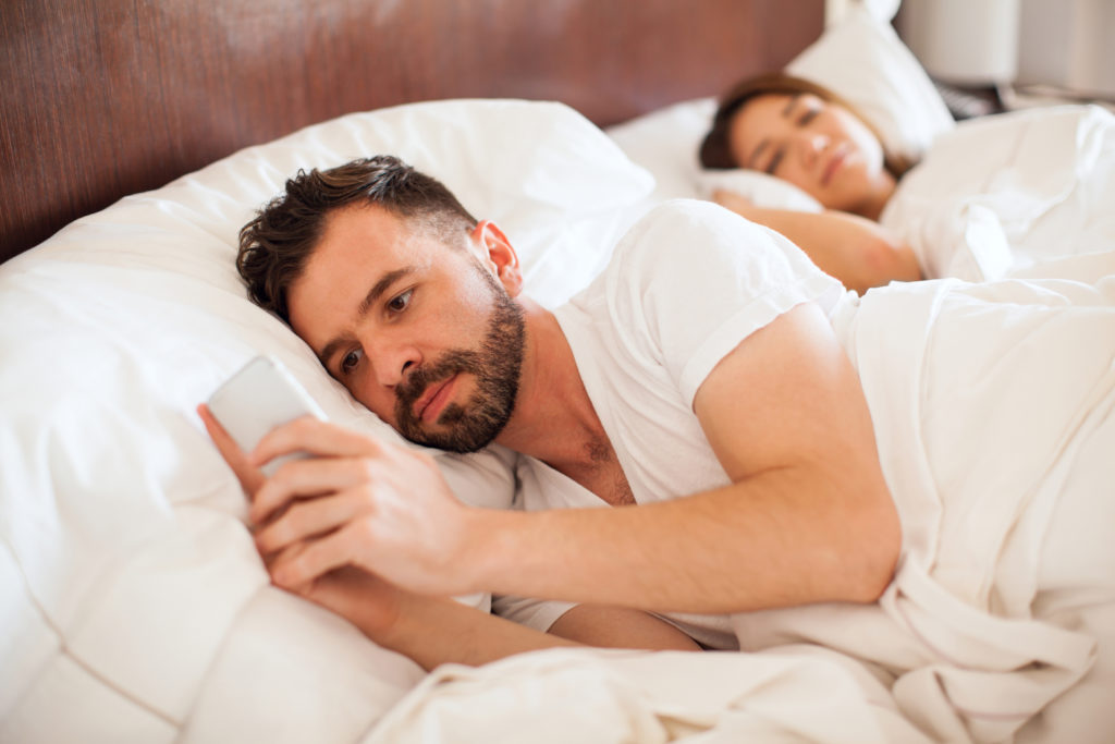 Portrait of a handsome young man with a beard checking his smartphone and texting his lover while his partner sleeps