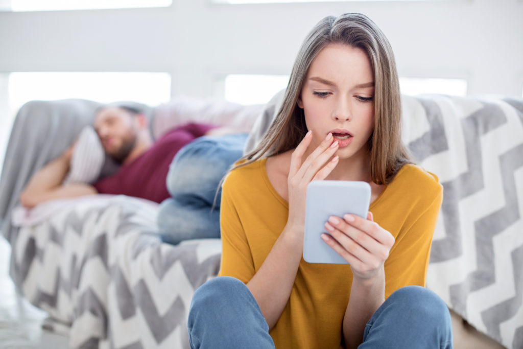 Oh no. Pretty perplexed long-haired young woman feeling surprised and holding her mans phone and he lying on the sofa