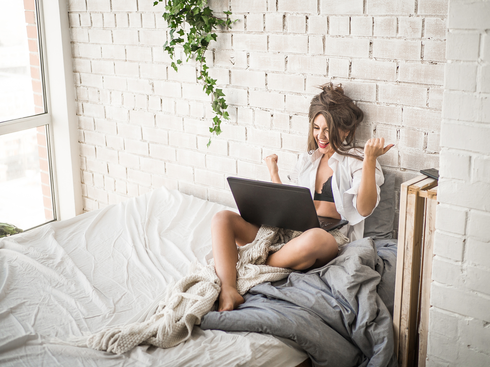 Happy casual beautiful woman working on a laptop sitting on the bed in the house.