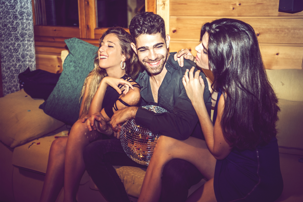 Two pretty women flirting with one man while sitting on sofa during private party.